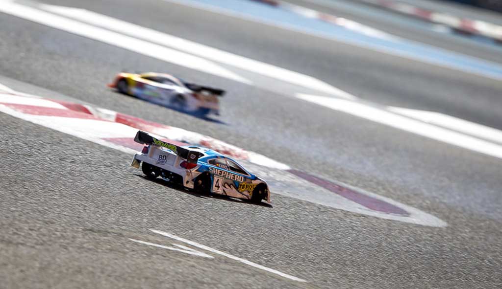 Dos coches RC touring de gasolina rodando sobre el asfalto de un circuito con pianos rojos y blancos
