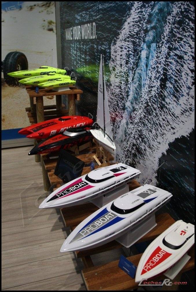 Varias lanchas de radiocontrol de casco blanco, rojo y verde junto a un velero, colocadas sobre soportes de madera en un expositor