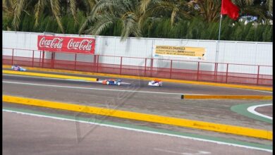 Coches de radiocontrol rodando en un circuito de asfalto con vallas publicitarias y palmeras al fondo durante una carrera