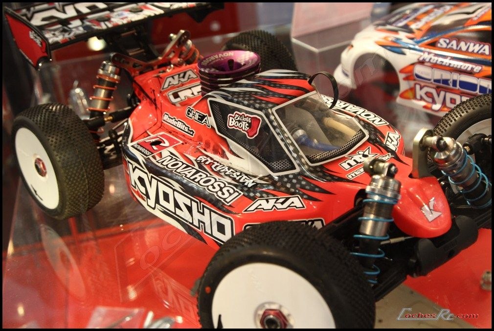 Buggy de radiocontrol de la marca Kyosho con carroceria roja y negra y logotipos del fabricante, expuesto sobre una peana