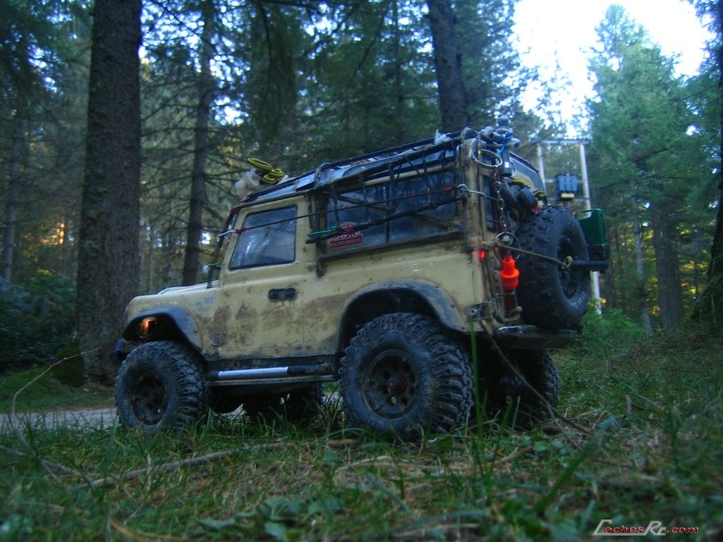 Crawler de radiocontrol con carroceria de todoterreno claro y rueda de repuesto, parado en un camino de bosque junto a troncos apilados